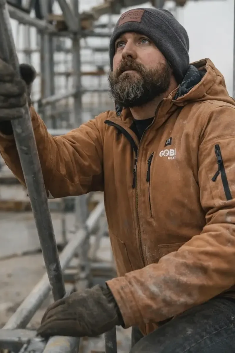 Tradesperson wearing camel heated work jacket kneeling on scaffolding with visible dust and tool belt contact during cold construction work.