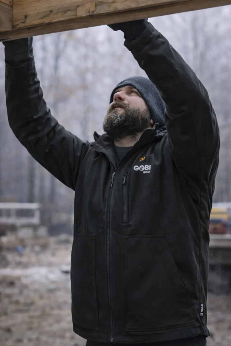 Tradesperson reaching overhead on a job site wearing the Grit heated jacket in onyx color, demonstrating mobility and range of motion during cold work
